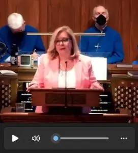 2022May-LindaLotz-TowsonPres-screencapture Linda Lotz standing at a lectern at Towson Presbyterian, with two members of the church choir in the background